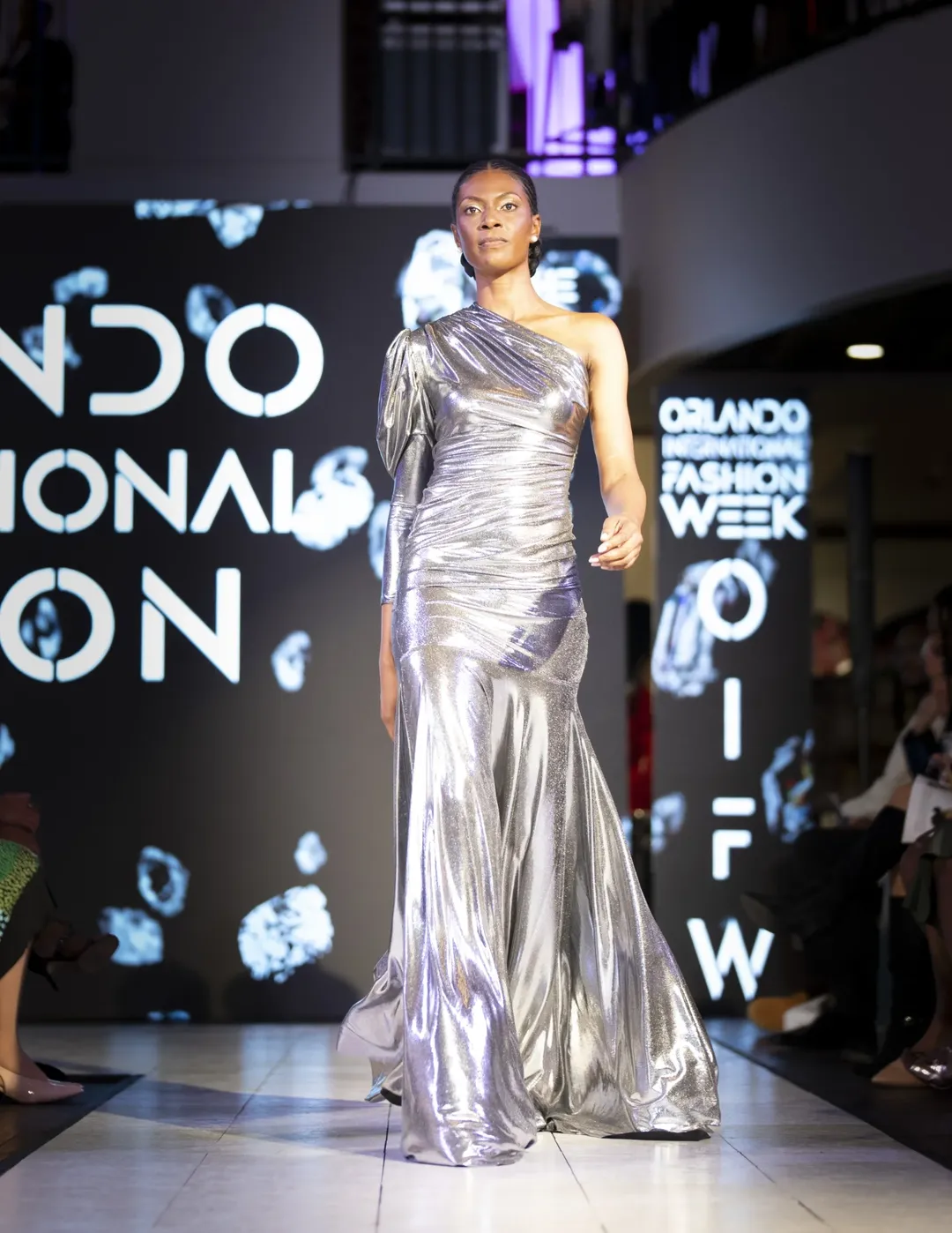 Model in a metallic gown walking a live runway with event branding and audience visible.