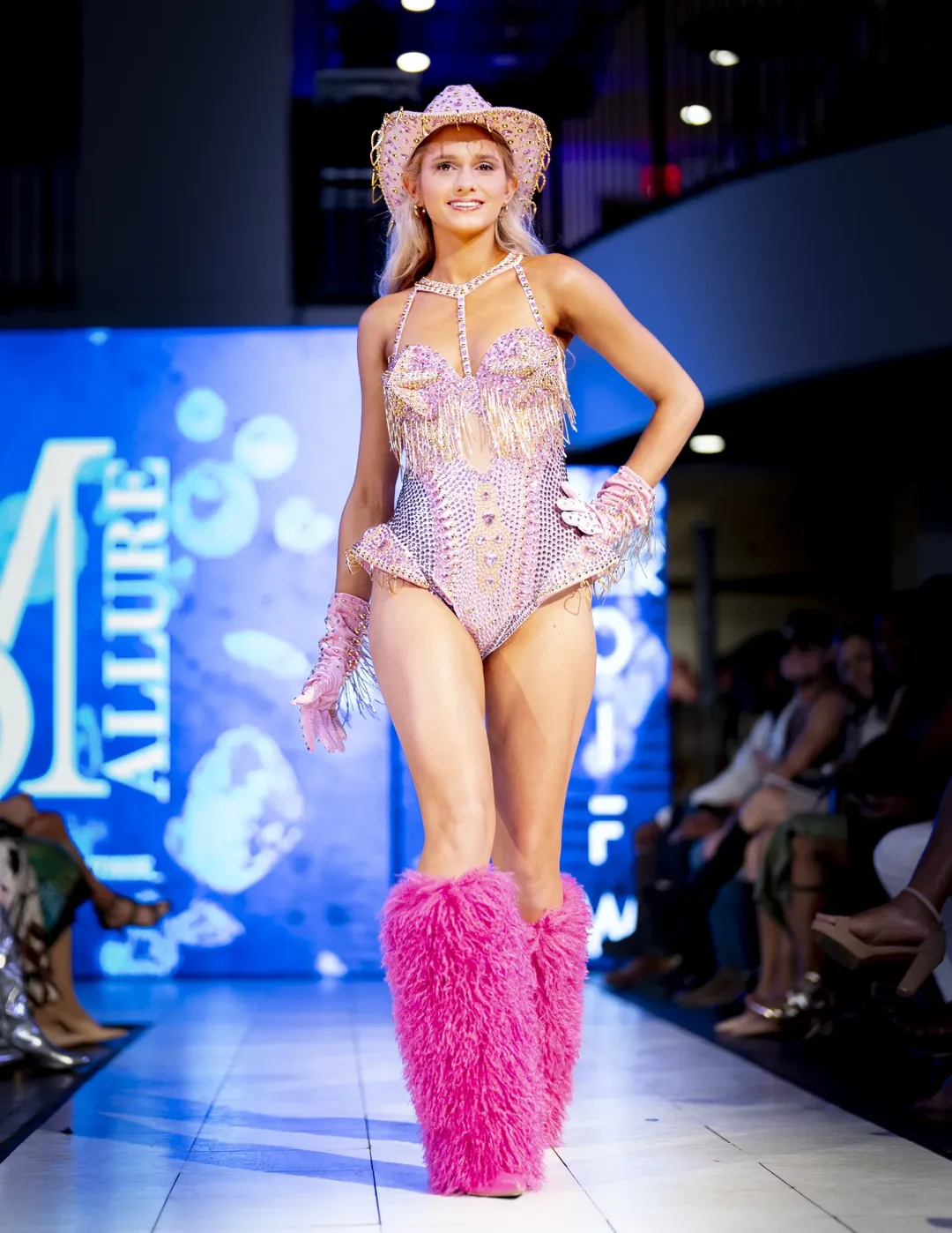 Model in a bright pink runway look at a live fashion event with stage lighting and audience visible.