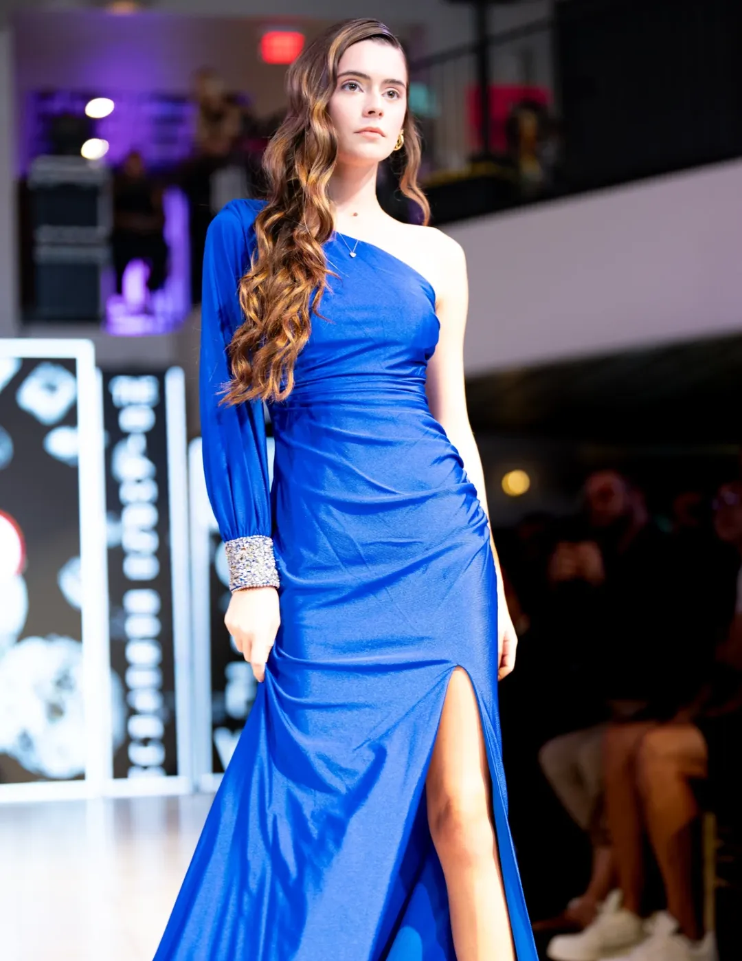 Model walking a brightly lit runway with audience seating visible at a live fashion event.