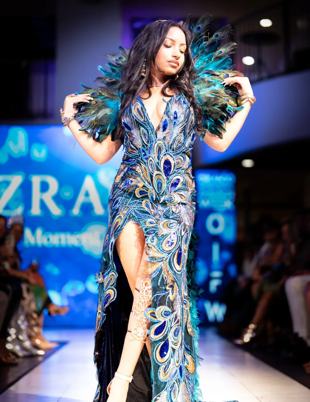 Model in a dramatic feathered runway look at a live event with stage lighting and audience visible.